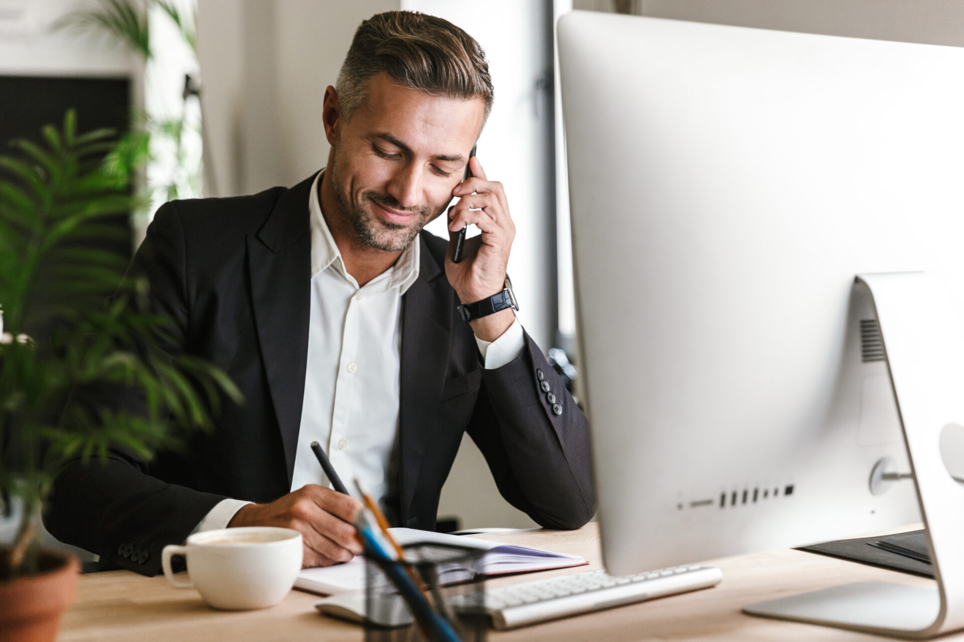 Image of handsome businessman talking on cell phone about remittance while working on computer in office